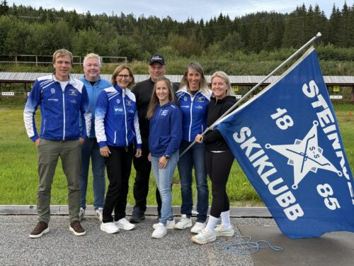 Gruppebilde av personer på et skiskytterstadion. holder et flagg med "Steinkjer skiklubb 1885"